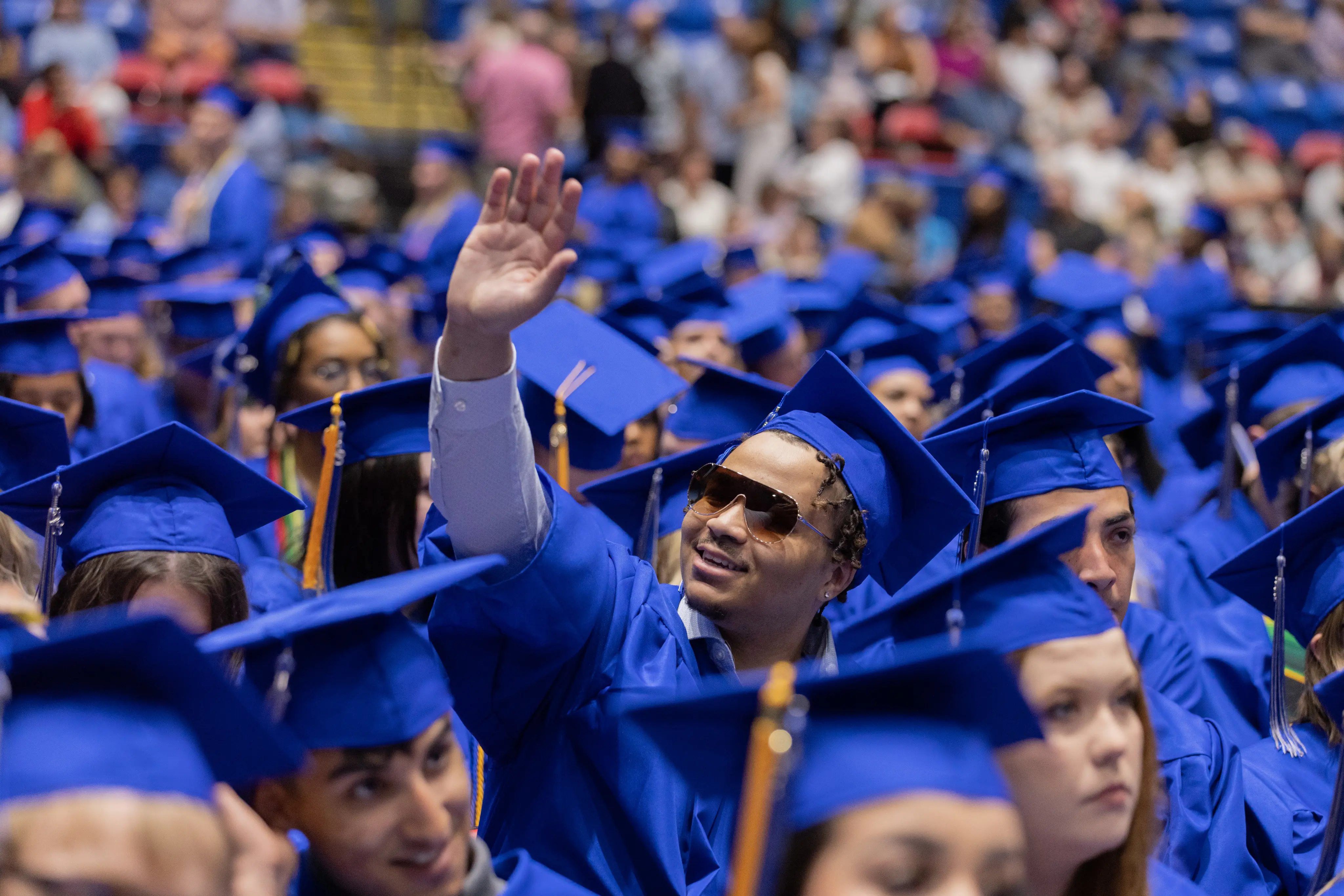 Graduate in blue cap and gown smiles and waves among a sea of fellow graduates during commencement.